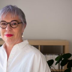 Andrea Pető looking into the camera, wearing black rounded glasses, red lipstick and a white shirt, with short grey hair, standing in front of a neutral background