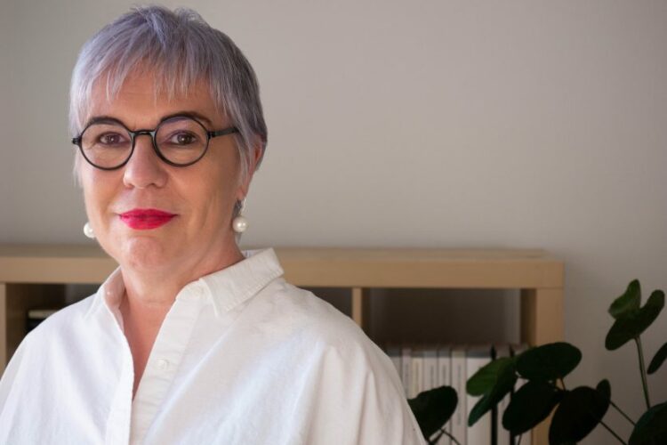 Andrea Pető looking into the camera, wearing black rounded glasses, red lipstick and a white shirt, with short grey hair, standing in front of a neutral background