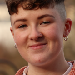 Luc O'Leary profile picture, looking into the camera, smiling, with short brown hair and shaved side of the head, wearing small circular earrings, a white t-shirt and an orange shirt.
