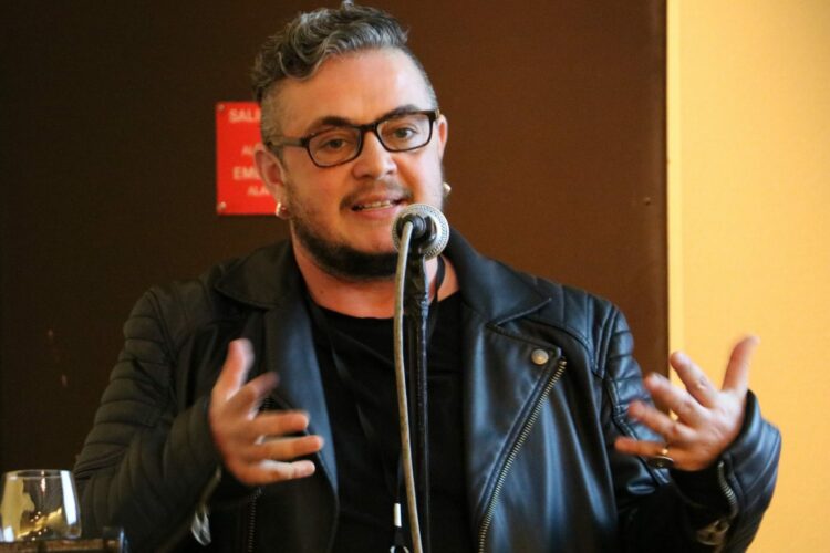 Mauro Cabral Grinspan speaking into a microphone wearing a black leather jacket, black-rimmed glasses and with short cropped black hair and beard