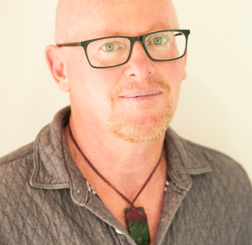 Jack Byrne headshot, looking into the camera wearing black-rimmed glasses, a black necklace and a brown t-shirt with a collar, bald head and ginger facial hair