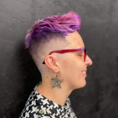Kot side portrait on a black background with pink spiky short hair, wearing pink glasses, a star-shaped metal earring and a black and white patterned top.