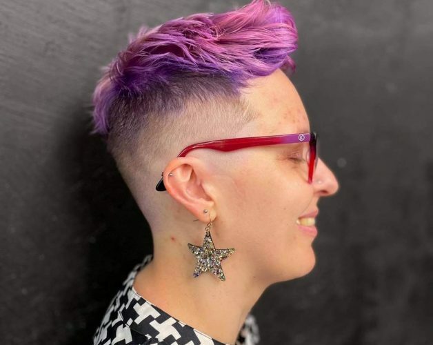 Kot side portrait on a black background with pink spiky short hair, wearing pink glasses, a star-shaped metal earring and a black and white patterned top.
