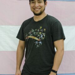 Michel Requelme standing in front of a trans flag, wearing a black t-shirt and a watch on his left wrist, smiling into the camera with short black hair and a cropped black beard