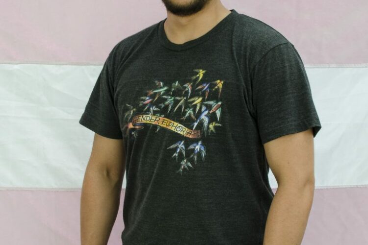 Michel Requelme standing in front of a trans flag, wearing a black t-shirt and a watch on his left wrist, smiling into the camera with short black hair and a cropped black beard