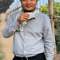 Kem Vichet standing outside looking straight into the camera, wearing black trousers, a light grey shirt, a white and grey checked scarf, with close cropped black hair.