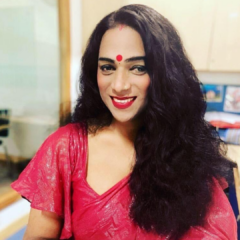 Abhina Aher sits on a chair in front of a mirror smiling, with long black wavy hair, wearing a pink sari, red lipstic and a bindi on her forehead.