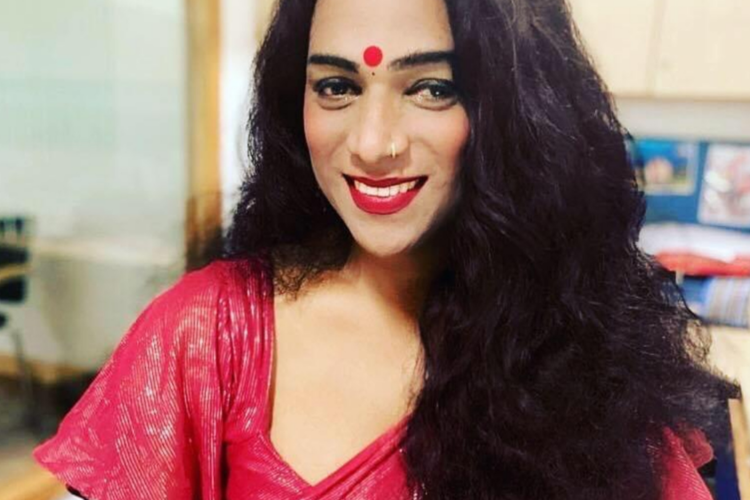 Abhina Aher sits on a chair in front of a mirror smiling, with long black wavy hair, wearing a pink sari, red lipstic and a bindi on her forehead.