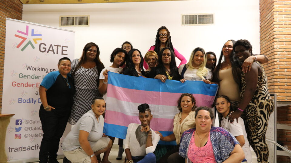 Group of trans activists from the Dominican Republic holding a trans flag and standing in front of a white background with GATE's logo