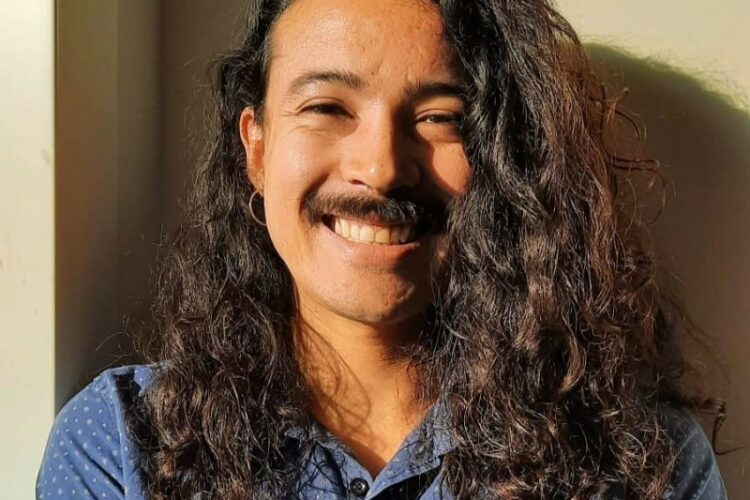 Douglas da Silva smiling into the camera against a cream background, with long dark curly hair and a dark mustache