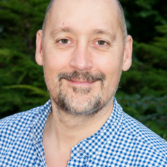 Morgan Carpenter smiling looking into the camera with short brown hair, a grey-black beard and wearing a white and blue checked shirt standing in front of greenery.