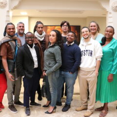 A group of 12 trans and gender diverse people of various ethnicities stand in a group in a large hallway with two white pillars behind them