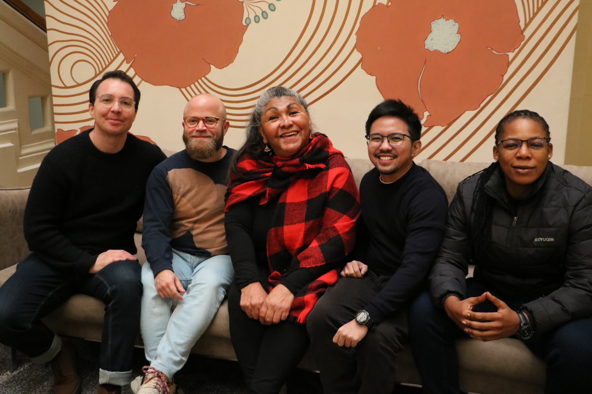 Five people sit side by side on a sofa, smiling at the camera against a patterned background.