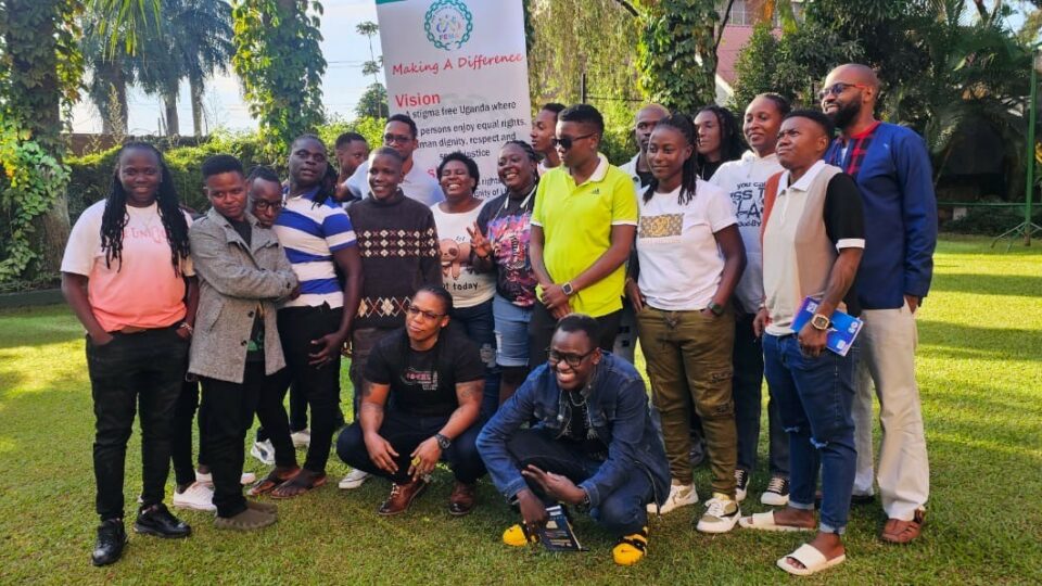 A group of 19 Ugandan trans activists standing outside on grass in the sunshine with the Fem Alliance Uganda banner in the background