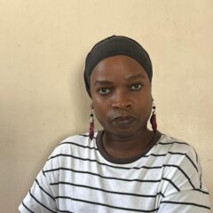 Headshot of an African trans woman wearing a white and black striped tshirt, a black headdress and red dangly earrings