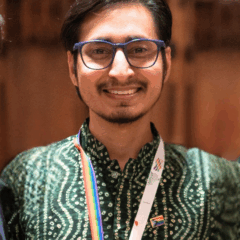 Photo of non-binary queer person from India smiling looking straight into the camera with dark brown hair parted on one side, a moustache and goatee, wearing blue glasses, a green and white patterned shirt and a rainbow flag-colored lanyard on a brown background.