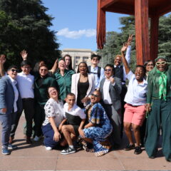 Group photo of Trans Advocacy Week delegates standing in front of the Broken Chair outside the UN Geneva, cheering and laughing