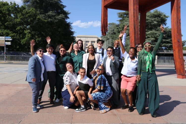 Group photo of Trans Advocacy Week delegates standing in front of the Broken Chair outside the UN Geneva, cheering and laughing